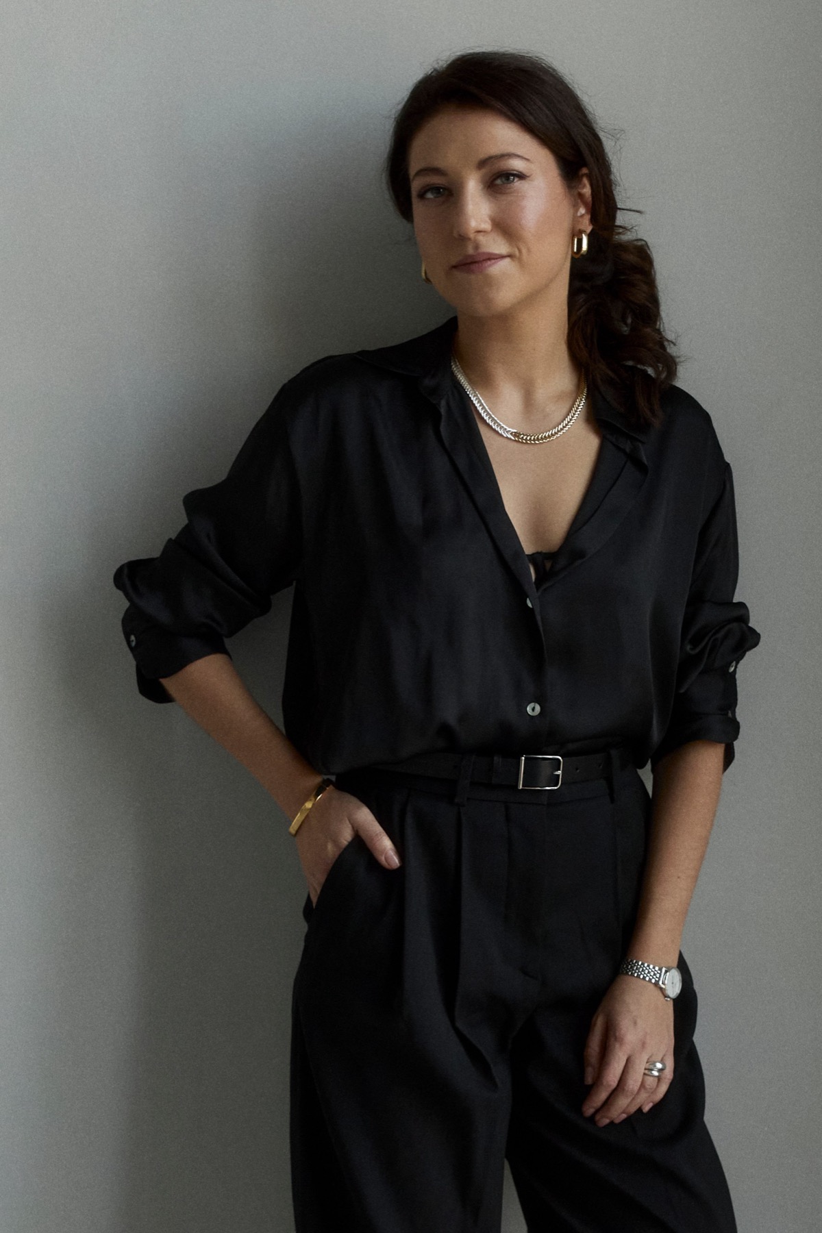 Ksenia in black, standing against a grey wall, looking at the camera