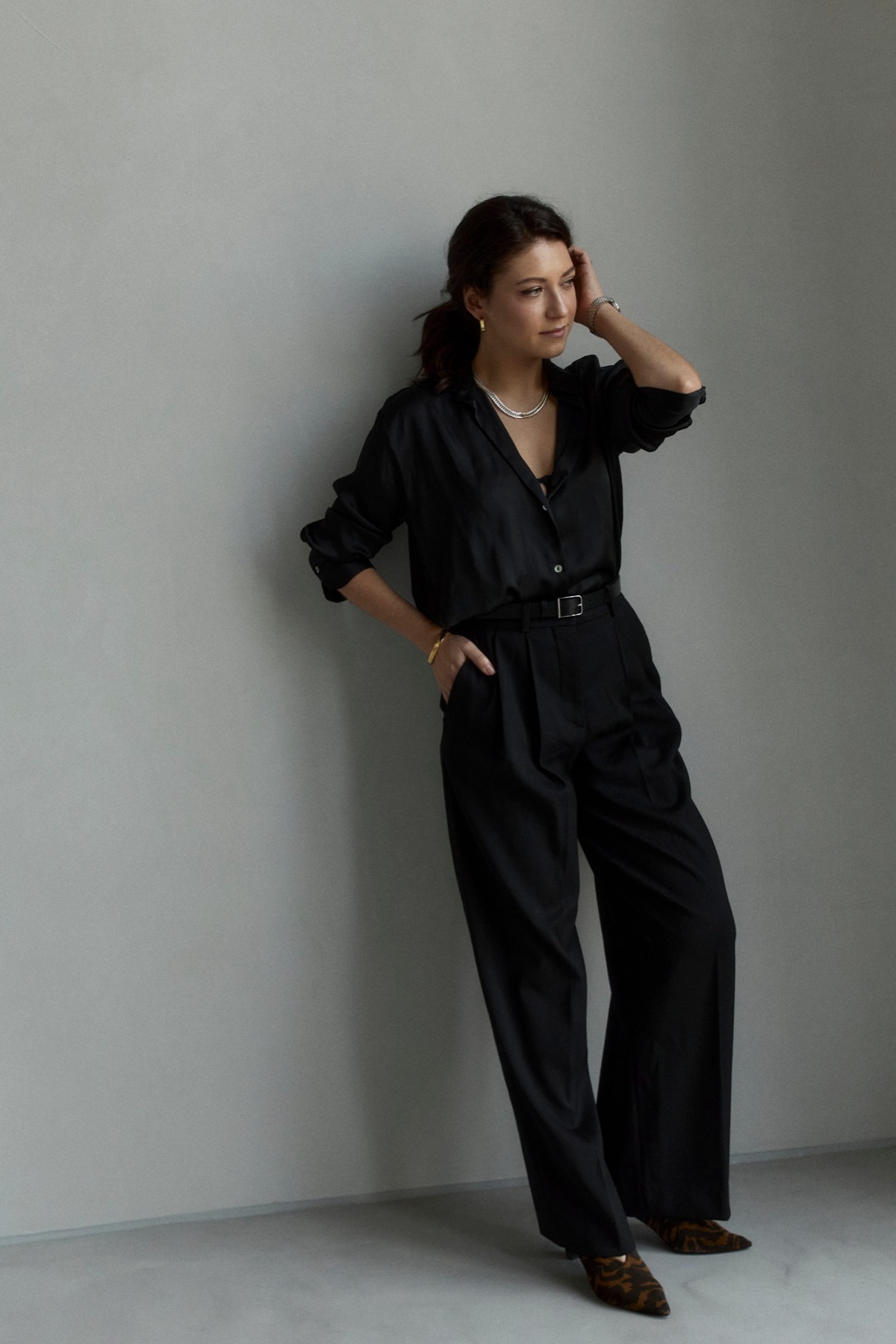 Ksenia in black, standing against a grey wall with one hand raised to her hair