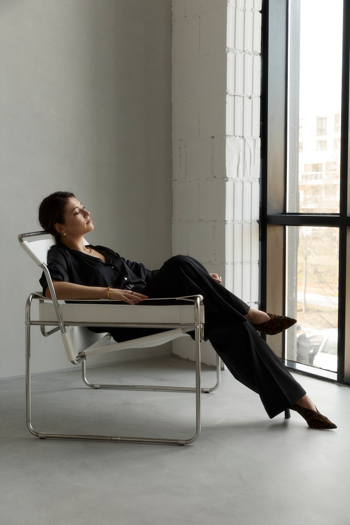 Ksenia reclining in a chrome chair beside a tall window and white brick column